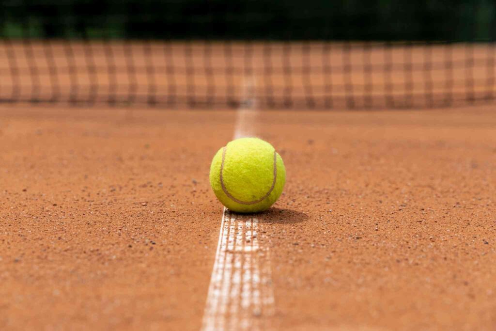 Bola tenis berwarna kuning tergeletak di lapangan tenis dengan garis putih yang terlihat jelas.