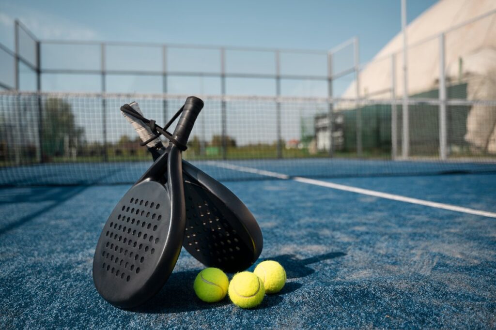 Dua raket padel dan beberapa bola tenis diletakkan di lapangan biru