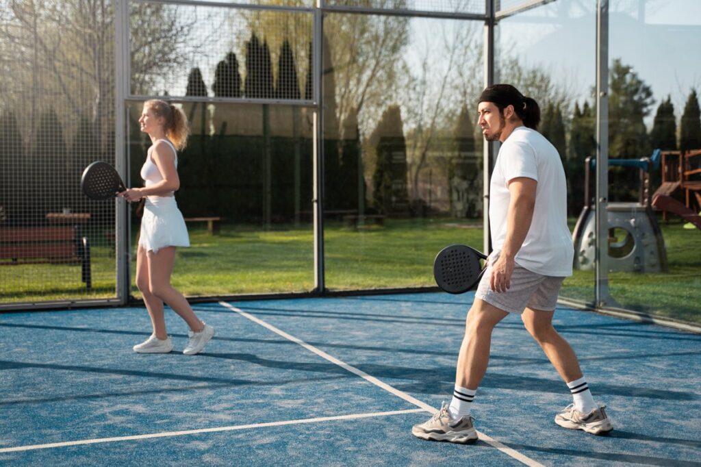 Dua pemain sedang bermain padel di lapangan outdoor berdinding kaca